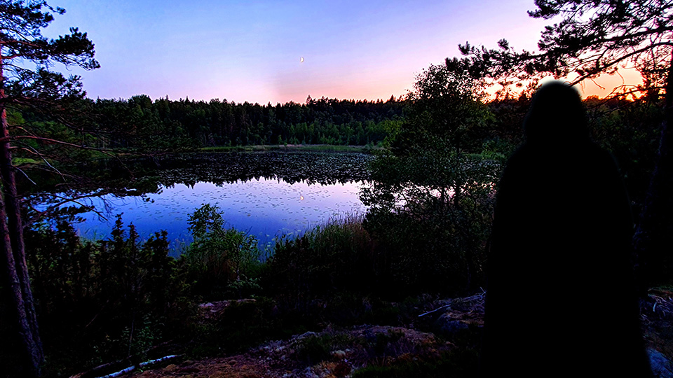 En mörk figur i kåpa blickar ut över en sjö i skymningen.