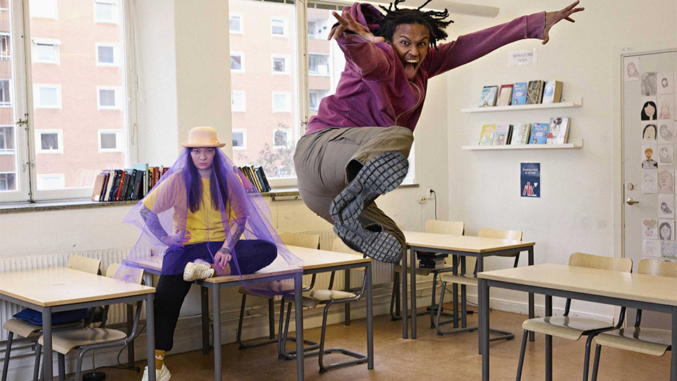 En ung kvinna med hatt och flor. Hon sitter i en skolbänk. En ung man i huvtröja hoppar högt i luften och ser ut att vråla mot kameran.