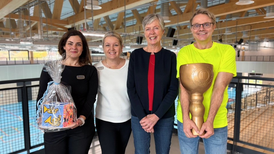 4 personer ler och kollar in i kameran. personen längst till vänster håller i ett föremål och personen längst till höger håller i en stor pokal.