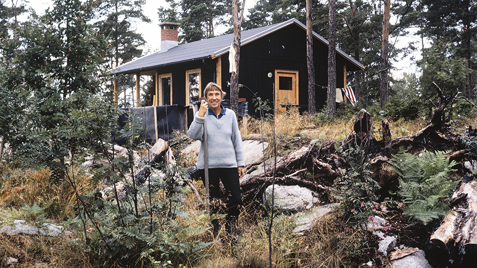 En glad kvinna i långbyxor och pullover står framför en låg timmerstuga.