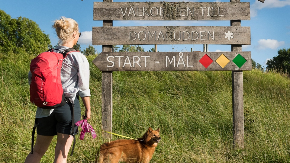 En kvinna med ryggsäck och en hund står framför en skylt.