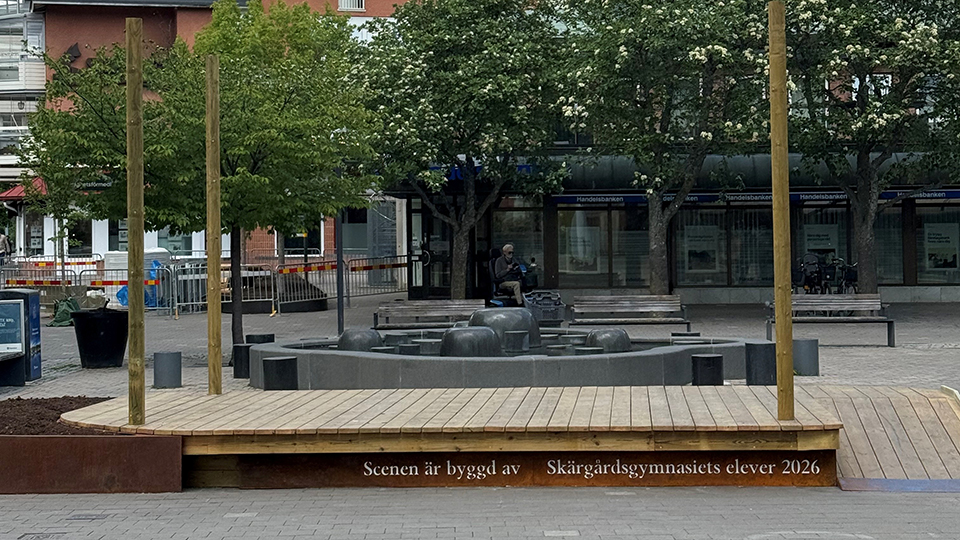 En scen gjord av trä på ett torg. Scenen har en skylt där det står "Scenen är byggd av Skägårdsgymnasiets elever 2026".