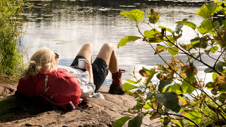 Kvinna i friluftskläder ligger och vilar på en klippa vid en sjö med en ryggsäck under huvudet.