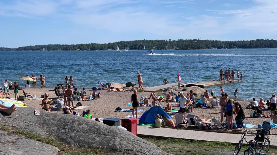 En solig sommardag på en sandstrand. Det är många människor i badkläder på stranden.