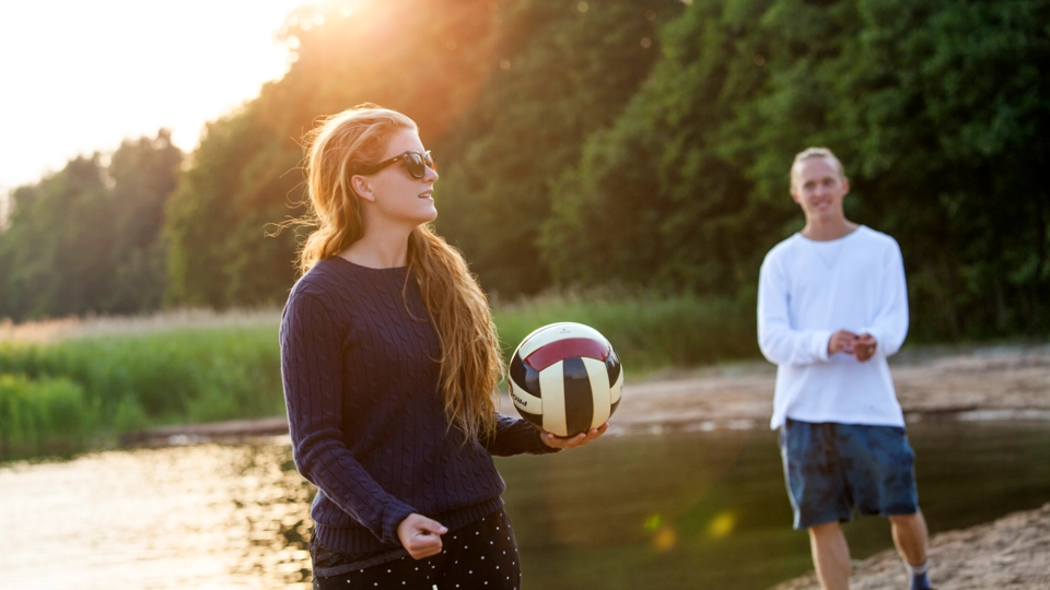 En kvinna med långt brunt hår och solglasögon håller i en volleyboll på en strand vid vattenbrynet. Bredvid henne står en man med en vit långärmad t-shirt och shorts. I bakgrunden kan man se vatten, vass och grönskande träd samtidigt som solen går ner över trädtopparna.