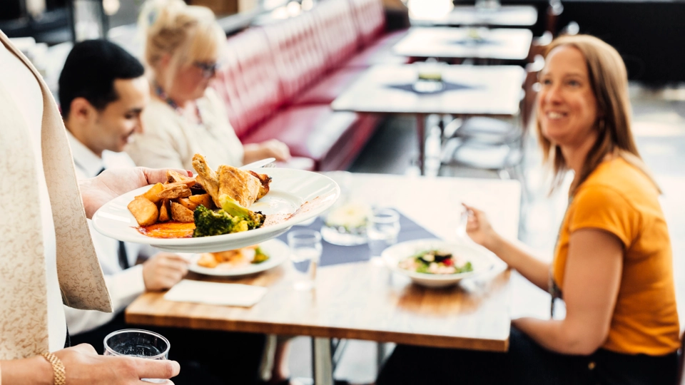 Några personer sitter vid ett matbord. En person står framför med en tallrik fylld med mat.