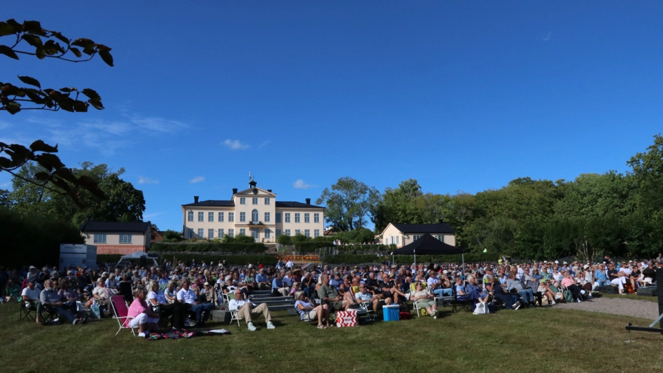 Bild på publiken i slottsparken med Östanå slott i bakgrunden. Himlen är blå och i förgrunden syns grönskande blad.