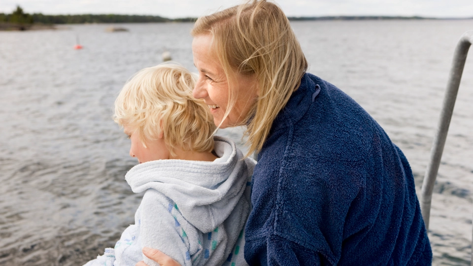 En äldre kvinna håller om en liten pojke och de sitter vid vattnet.
