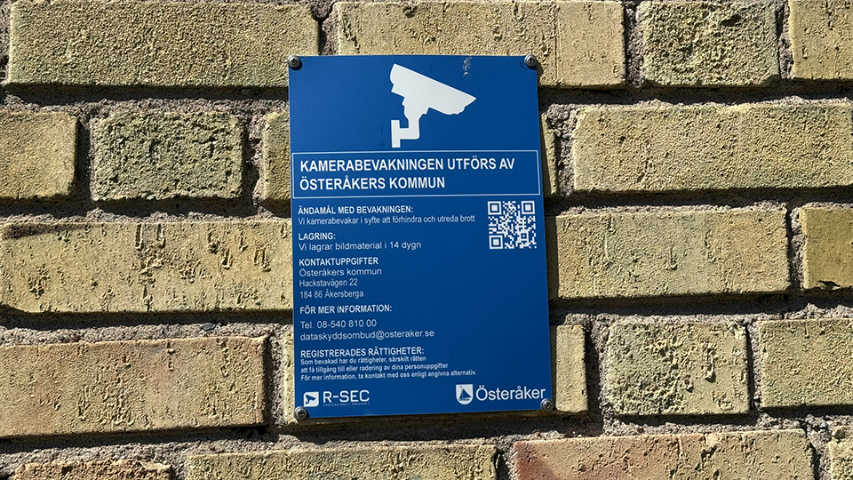 Skylt för kamerabevakning från Österåkers kommun sitter på gul tegelvägg