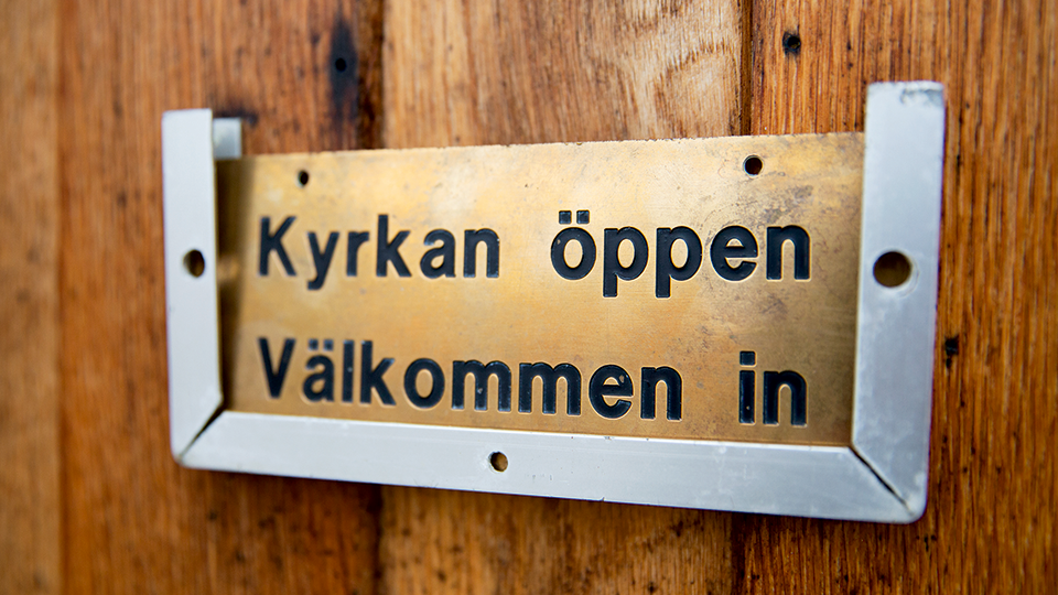 En mässingsskylt med texten "Kyrkan öppen" sitter på en dörr i trä.