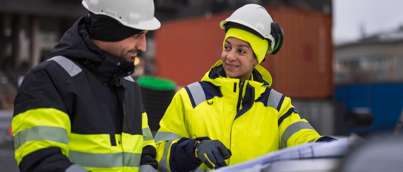 En kvinna och en man står i varselkläder och blickar ut över en stor ritning. Båda har vita bygghjälmar på huvudet och i bakgrunden kan man skymta containrar.