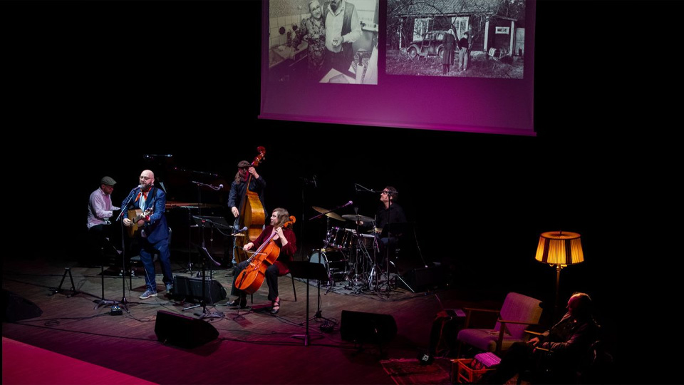 En bild på en scen. Där står ett band med cello, bas, gitarr, piano och slagverk. Vid sidan står två djupa fåtöljer.