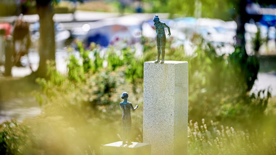 Två pelare i betong. På den högre står en figur i brons och tittar ned.