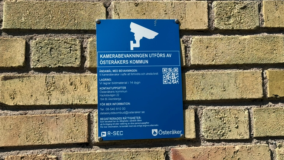 En tegelvägg med en blå skylt där det finns information om kamerabevakning som utförs av Österåkers kommun.