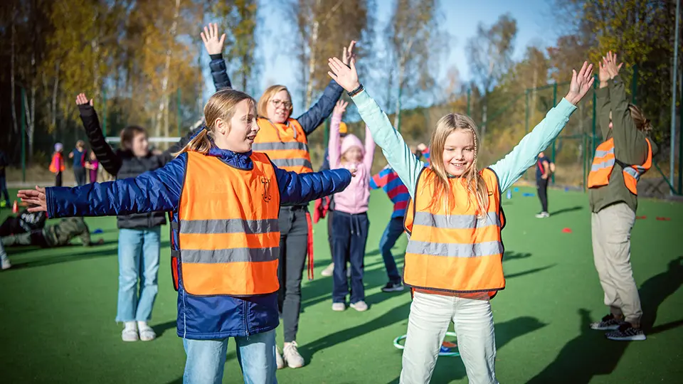 Bilden visar barn som har reflexvästar i orange på sig. Barnen leker tillsammans på en forbollsplan.