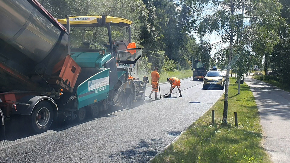 Beläggningsarbeten på en väg med en stor asfaltsmaskin och två personer i orange varselkläder.