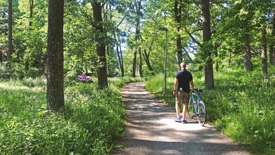 En man leder sin cykel på en grusad promenadväg omgiven av grönskande träd och växter längs Åkers kanal.