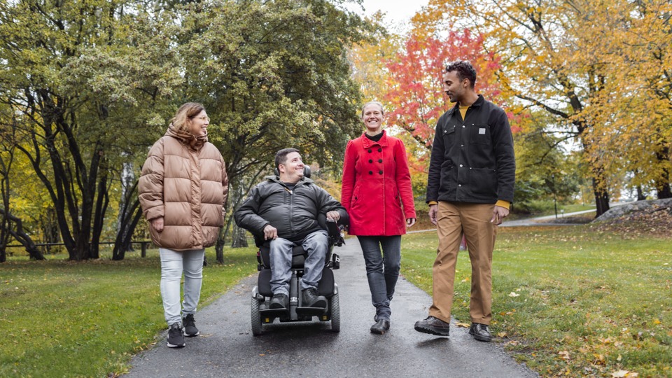 Fyra personer går i en park, en av personerna är rullstolsburen. Alla ser glada ut.