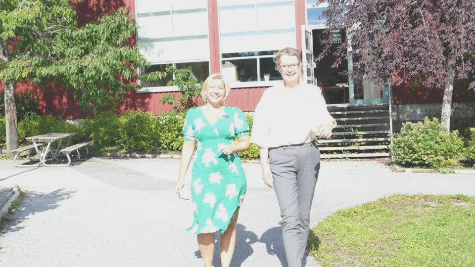 Rektor Jenny Nyrén och skolminister Lina Axelsson Kihlblom går sida vid sida utomhus på skolgården med Skärgårdsstadsskolans röda skolbyggnad i bakgrunden. Solen skiner och trädens blad är gröna. Det är sommar och båda kvinnorna ser glada ut.