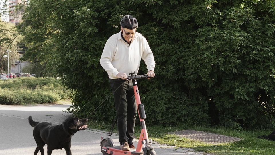 Man åker elsparkcykel när han rastar sin hund.
