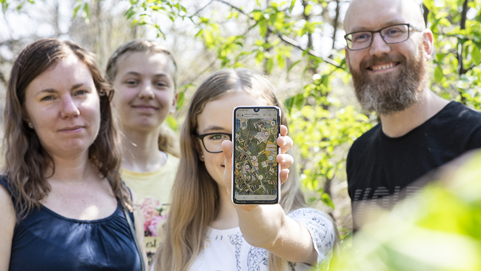 Familj ståt på rad. Yngsta dottern står i mitten och håller fram en mobiltelefon. På skärmen syns appen för Hittaut.