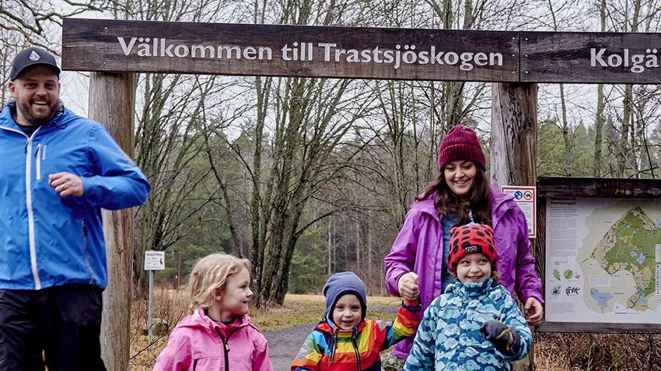 Bilden visar två vuxna och tre barn som springer och skrattar utomhus i färgglada ytterkläder. Det är höst och de spriger under en port av trä där det står Välkommen till Trastsjöskogen