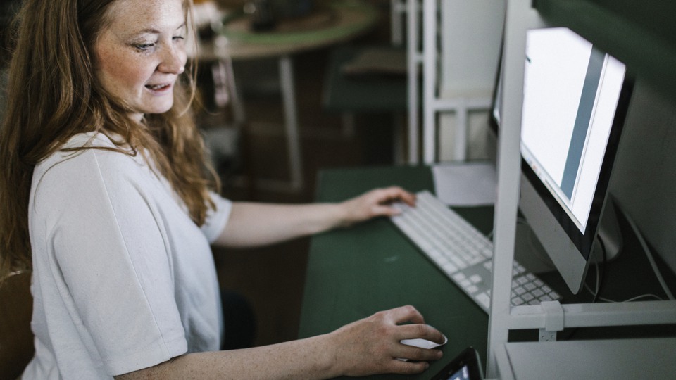 Person sitter vid ett skrivbord med en dator som lyser upp. Personen bär en vit tröja