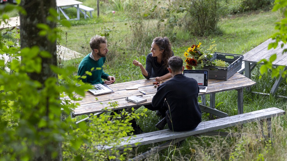 Tre personer sitter i möte vid ett bord utomhus. Det är grönt omkring och på bordet finns datorer, block och pennor