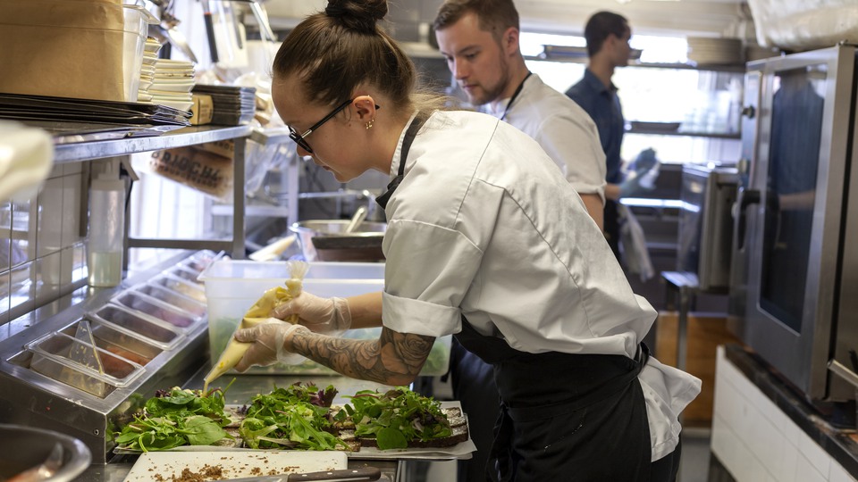 Tre personer i ett restaurangkök. Personen längst fram i bild spritsar sås på sallad 