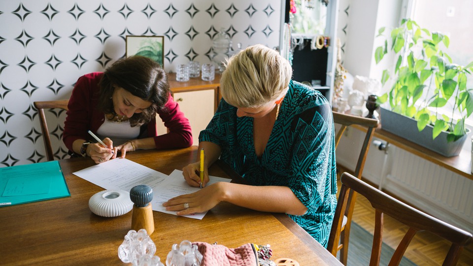 Två personer sitter vid köksbord i hemmamiljö, skriver eller fyller i papper