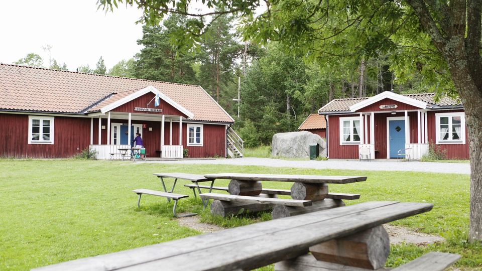 Två röda stugor vid skog. På gräsmatta framför husen står flera bänkar och bord i trä