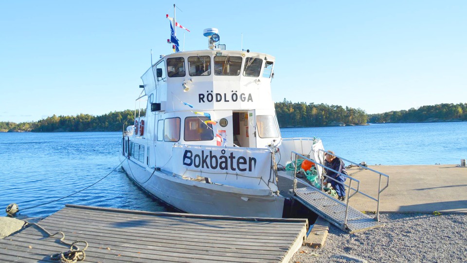 Bilden visar Bokbåten Rödlöga som lagt till vid en kajplats. Det är blå himmel och soligt.
