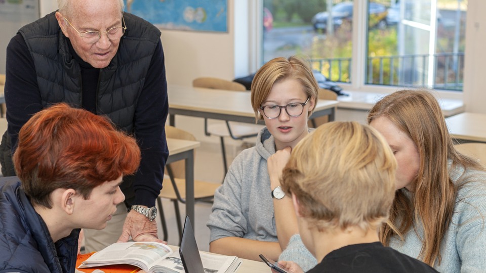 Bilden vsar en lärare och elever som sitter vid ett skrivbord i ett klassrum