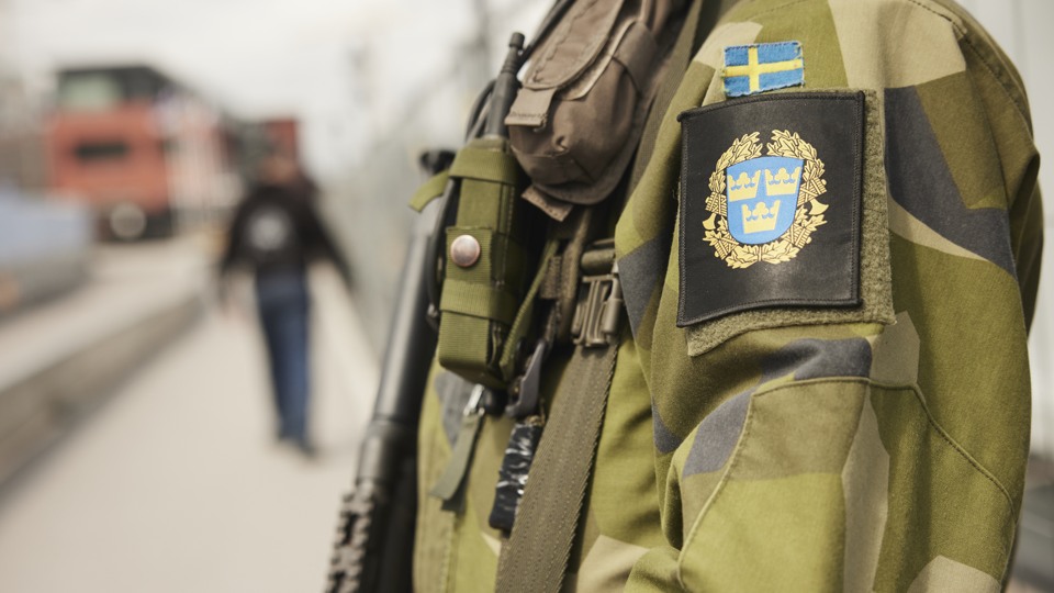 Bilden visar skjortärmen på en militär i uniform. I bakgrunden kan man ana att det är någon annan som går. Det ser ut som att det är en tågperrong.