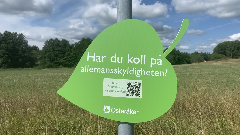 En grön skylt i form av ett löv sitter på en lykstolpe vid ett sädesfält. På skylten står det "har du koll på allemansskyldigheten?"