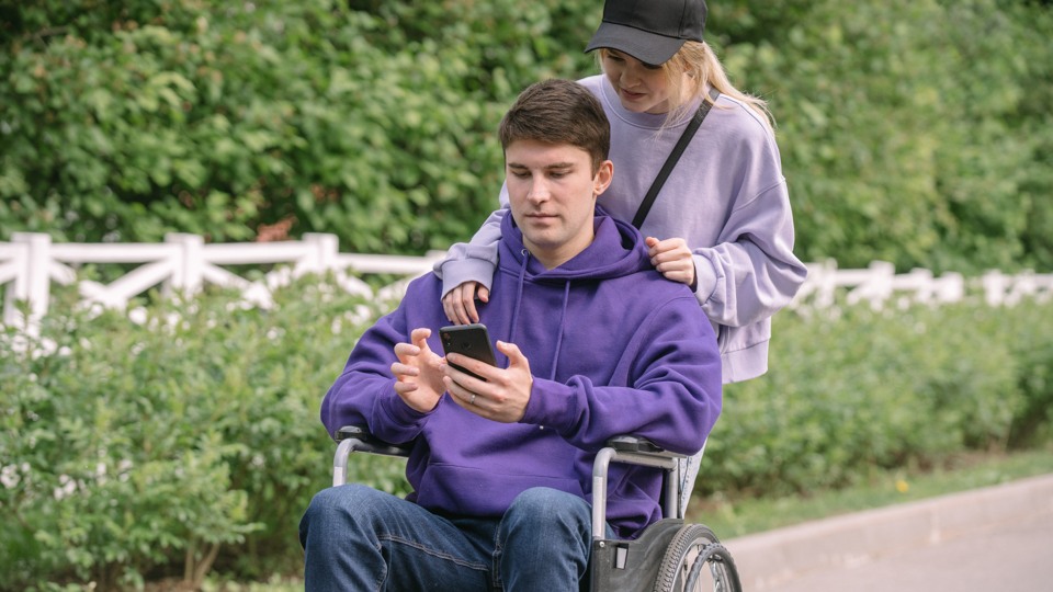 Man i rullstol som har mobiltelefon i handen. Kvinna står bakom rullstolen.