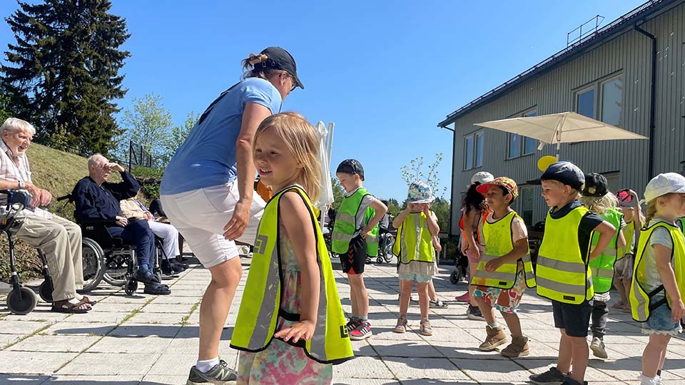 Bilden visar en solig vår dag. Ute på en innergård sitter pensionärerna från ett äldreboende och tittar på medans förskolebarn har dansuppvisning.