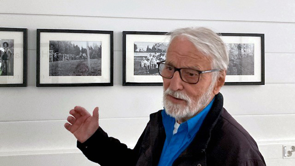 En äldre herre framför en vägg med svart-vita fotografier.