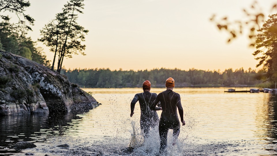 Två person i våtdräkt och badmössa springer i vattnet. De syns bakifrån och i solnedgång. 