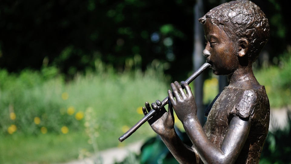 Konstverk  av Lena Lervik. En figur i brons föreställande en pojke som spelar flöjt. 