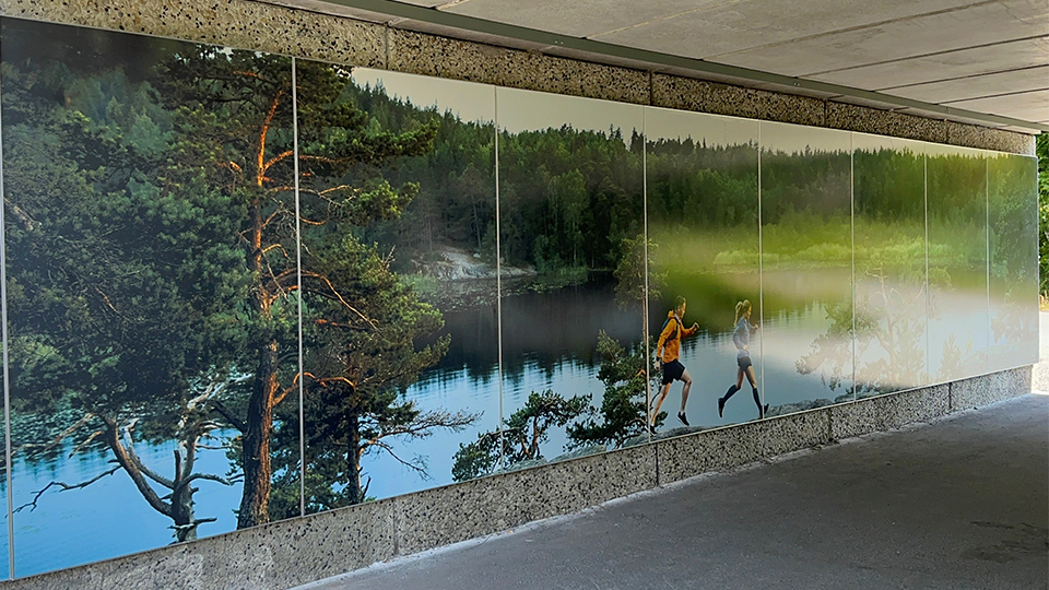 Gångtunnel i betong. På väggen sitter ett fotografi som visar en skogig miljö. Två personer i sportkläder springer på toppen av ett berg med en sjö i bakgrunden. Det är sommar.