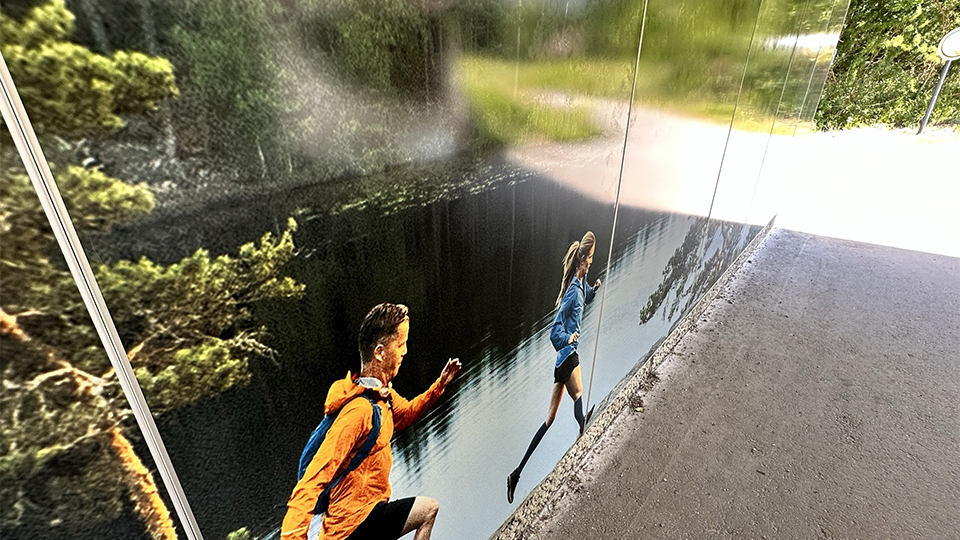 Närbild ur fotoväggen som visar de två personerna i sportkläder som springer. Det är en solig dag och gångvägen speglar sig i den blanka väggen.