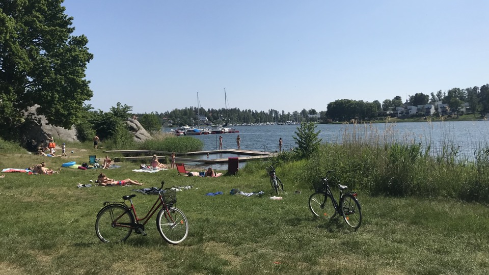 Badplats med gräs och sand. Badbrygga går ut i vattnet. Det ligger människor och solar. cyklar är parkerade vid sidan av.