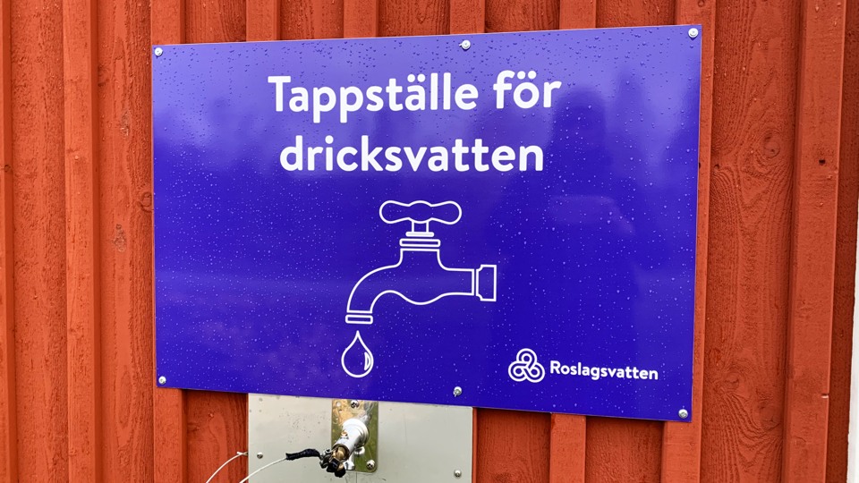 En lila skylt med texten Tappställe för dricksvatten och en bild av en kran sitter på en faluröd fasad.