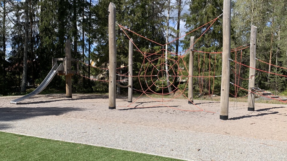 En klätterställning i trä med rep i formen av ett spindelnät. Intill syns en rutschkana. I förgrunden en konstgräsplan.