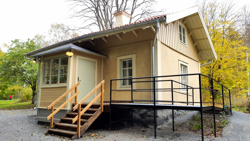 Blekgult mindre hus med veranda. Det är putsat. En tillgänglighetsanpassad ramp leder upp till verandan och entrén