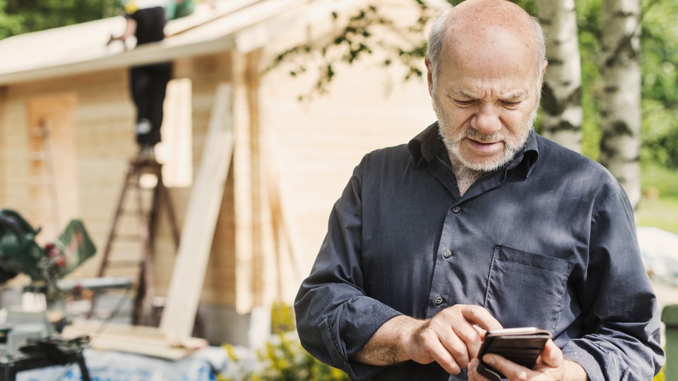 En man som står framför ett husbygge och tittar i sin mobiltelefon.