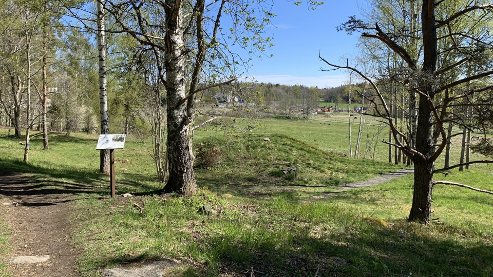 Grönskande omgivningar och stig. En kulle med kollapsad mitt syns till höger i bild. Framför står en informationsskylt.