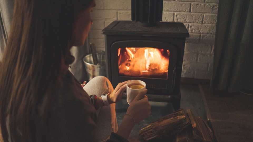 En kvinna sitter framför en kamin som brinner och dricker en kopp choklad
