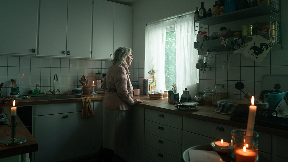 Bilden visar en äldre dam som står och tittar ut genom sitt köksfönster. I köket är det mörkt, elen fungerar inte och stearinljus är tända.